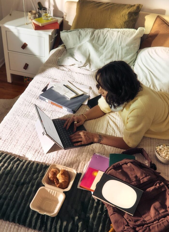 Woman working on laptop on bed with snacks and books.